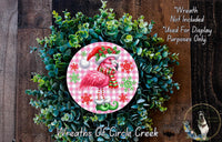 A wreath made of green foliage and featuring a pink flamingo wearing a Santa hat and scarf.