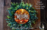 a wreath with a welcome sign in the center, surrounded by various fall-themed elements such as sunflowers, pumpkins, and pine cones. The wreath is displayed on a wooden surface, and there is a small dog in the bottom right corner of the image.