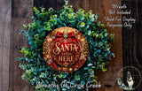 A wreath made of green leaves and a red ribbon with the text "Santa stops here" in the center, placed on a wooden surface.