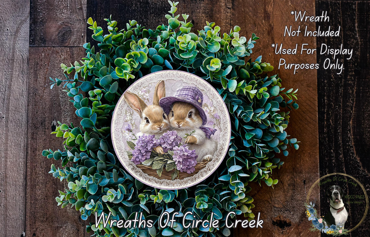 A wreath featuring two rabbits wearing purple hats, surrounded by green foliage. The wreath is displayed on a wooden surface, and there is a text overlay that reads "Wreath Not Included * Used For Display Purposes Only".