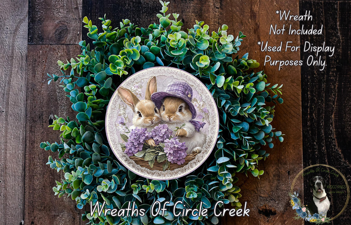 A wreath featuring two rabbits wearing purple hats, surrounded by green foliage. The wreath is displayed on a wooden surface, and there is a text overlay that reads "Wreath Not Included * Used For Display Purposes Only".