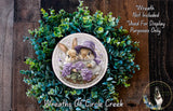 A wreath featuring two rabbits wearing purple hats, surrounded by green foliage. The wreath is displayed on a wooden surface, and there is a text overlay that reads "Wreath Not Included * Used For Display Purposes Only".