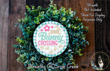 a wreath made of green leaves and flowers, with a circular sign in the center that says "Bunny Crossing". The sign has a pink flower and a brown rabbit on it. The wreath is displayed on a wooden surface, and there is a small dog in the bottom right corner of the image.
