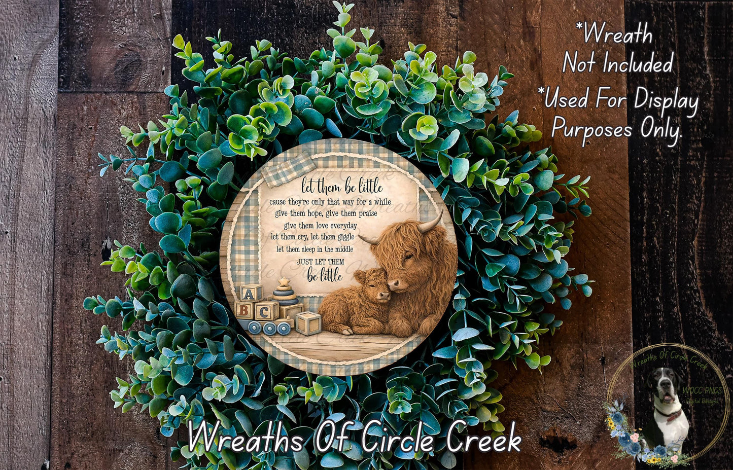 A wreath made of green leaves and a wooden sign with a mother cow and her baby, surrounded by text that reads "Let them be little. cause they're only that way for a while. give them hope, give them love, give them family. let them grow up to be just like you. let them sleep in the middle. let them be little." The wreath is displayed on a wooden surface, and there is a small dog in the bottom right corner of the image.