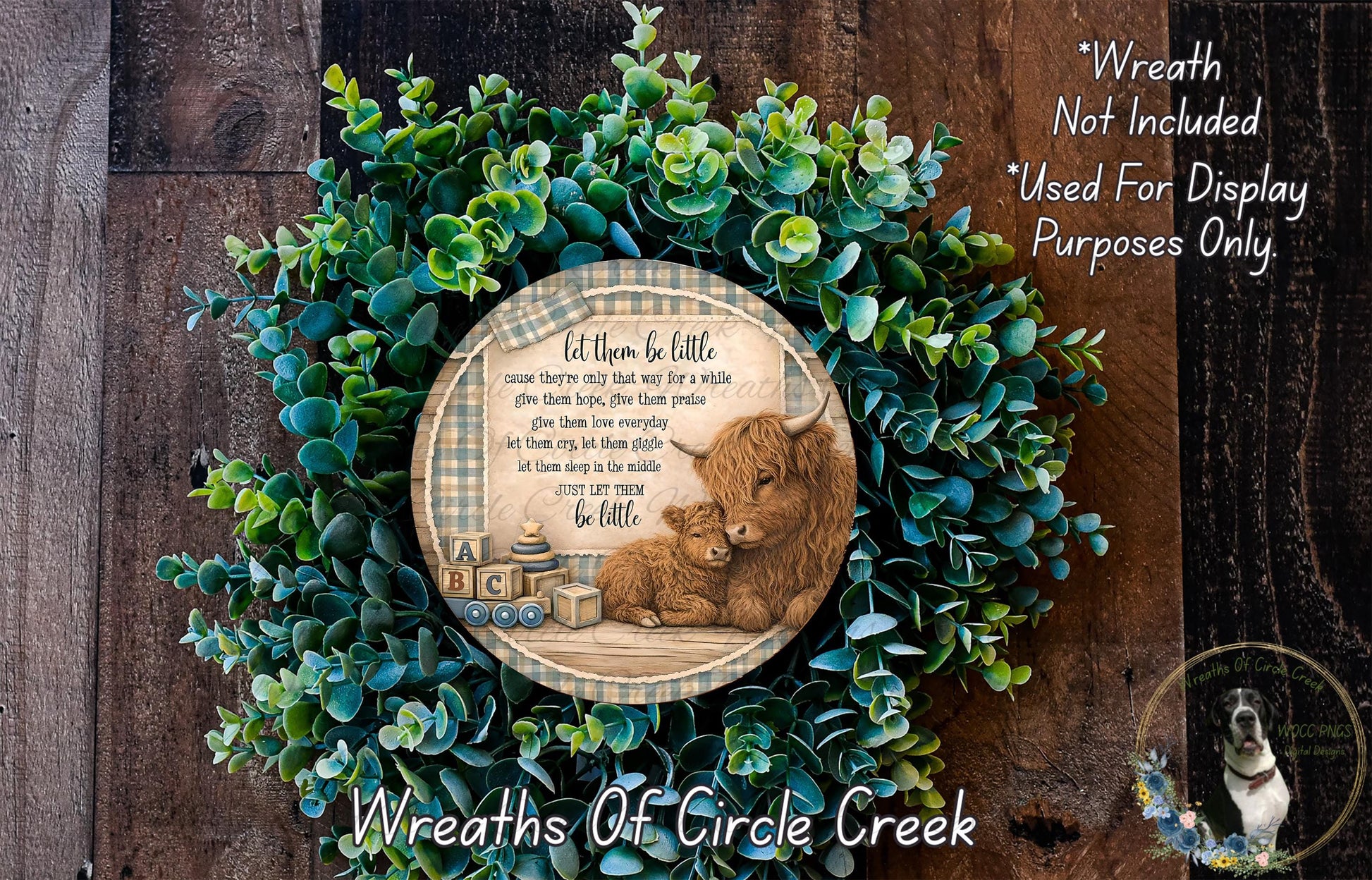A wreath made of green leaves and a wooden sign with a mother cow and her baby, surrounded by text that reads "Let them be little. cause they're only that way for a while. give them hope, give them love, give them family. let them grow up to be just like you. let them sleep in the middle. let them be little." The wreath is displayed on a wooden surface, and there is a small dog in the bottom right corner of the image.