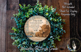 A wreath made of green leaves and a wooden sign with a mother cow and her baby, surrounded by text that reads "Let them be little. cause they're only that way for a while. give them hope, give them love, give them family. let them grow up to be just like you. let them sleep in the middle. let them be little." The wreath is displayed on a wooden surface, and there is a small dog in the bottom right corner of the image.