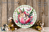 A decorative plate featuring a sleeping bunny nestled in a pink tulip, surrounded by green foliage and flowers, placed on a wooden surface.