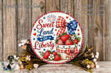 a round wooden sign with a patriotic design, surrounded by flowers and a basket of strawberries. The sign reads "Sweet Land of Liberty" and includes a fireworks display. The background is a wooden wall with a text overlay that says "Flowers Not Included Used For Display Purposes Only".