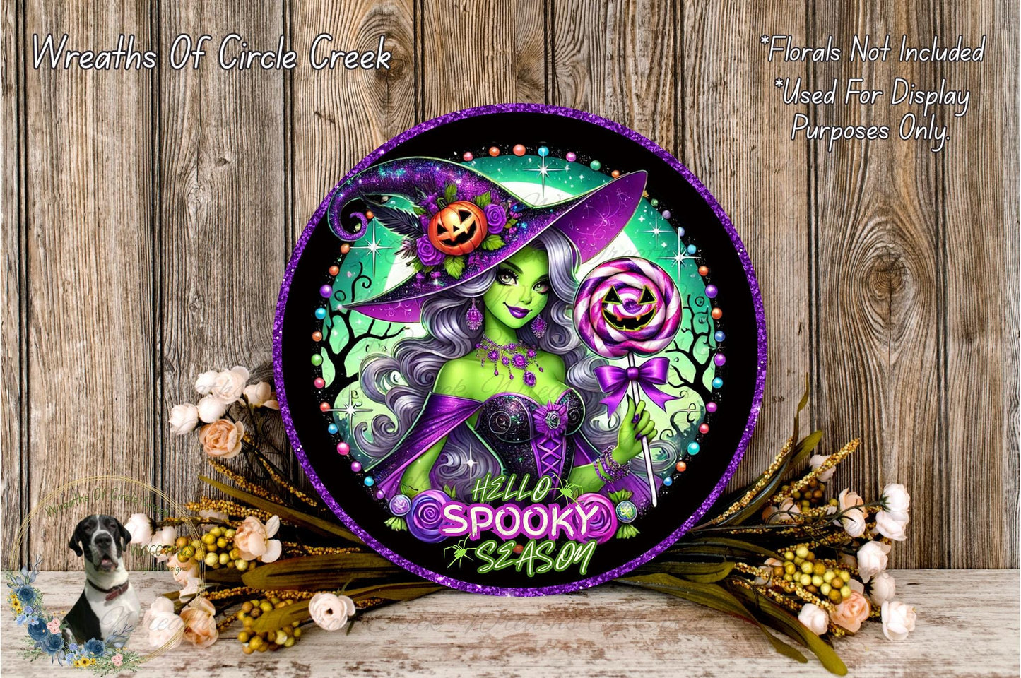 A decorative Halloween-themed wreath featuring a green-skinned witch with purple hair, holding a jack-o-lantern and a lollipop, surrounded by flowers and a wooden background.