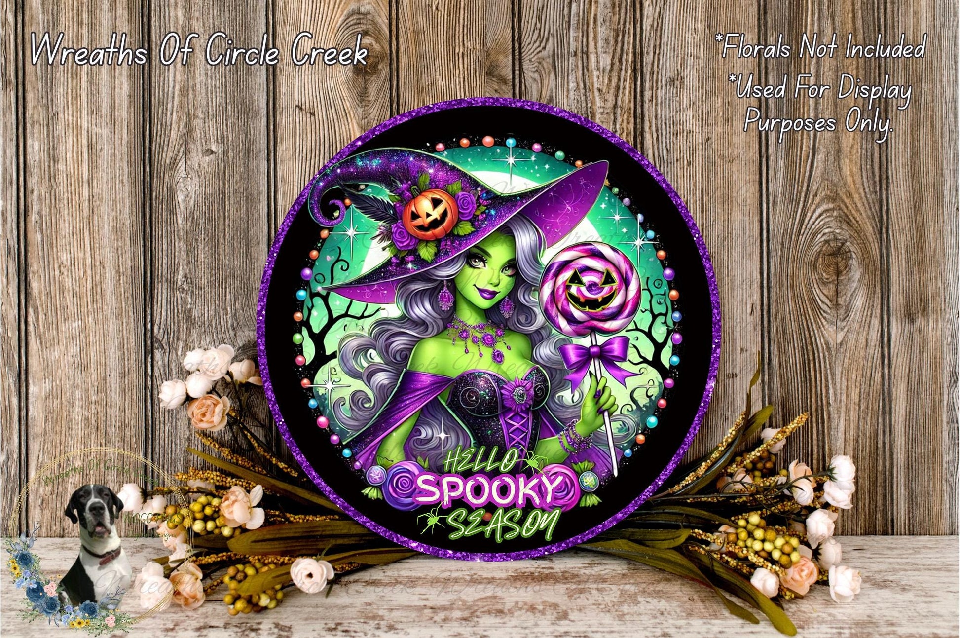 A decorative Halloween-themed wreath featuring a green-skinned witch with purple hair, holding a jack-o-lantern and a lollipop, surrounded by flowers and a wooden background.