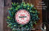 a wreath made of green leaves and flowers, with a red and white circular sign that reads "Happy Valentine's Day" in the center. The wreath is displayed on a wooden surface, and there is a small dog visible in the bottom right corner of the image.