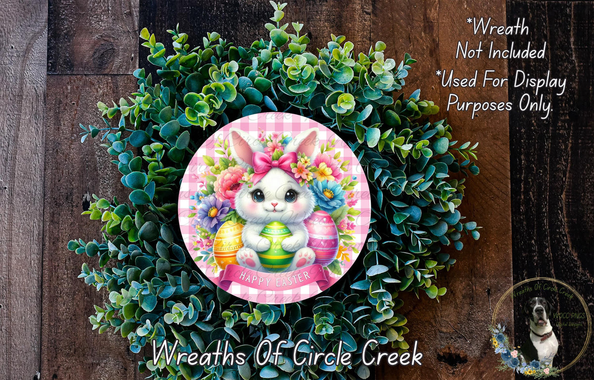 a wreath with a bunny wearing a pink bow and surrounded by green leaves and flowers. The wreath is placed on a wooden surface.