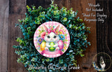a wreath with a bunny wearing a pink bow and surrounded by green leaves and flowers. The wreath is placed on a wooden surface.