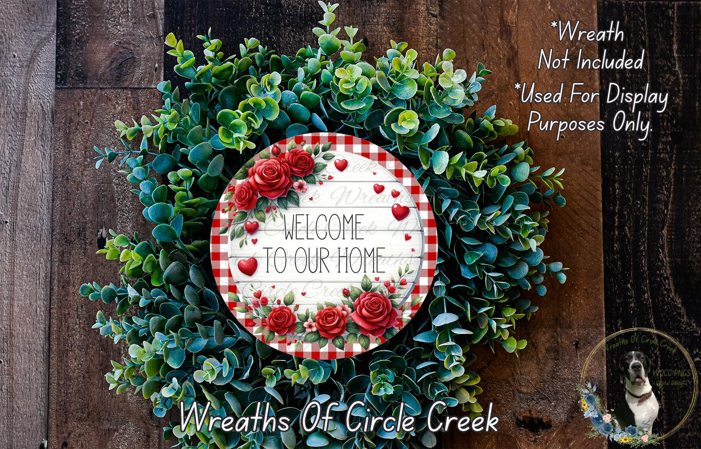 a wreath made of green leaves and red roses, with a sign that reads "Welcome to our home" in the center. The wreath is displayed on a wooden surface, and there is a small dog in the bottom right corner of the image.