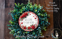 a wreath made of green leaves and red roses, with a sign that reads "Welcome to our home" in the center. The wreath is displayed on a wooden surface, and there is a small dog in the bottom right corner of the image.