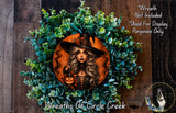 a wreath with a woman wearing a witch's hat and holding a lantern, surrounded by green foliage.