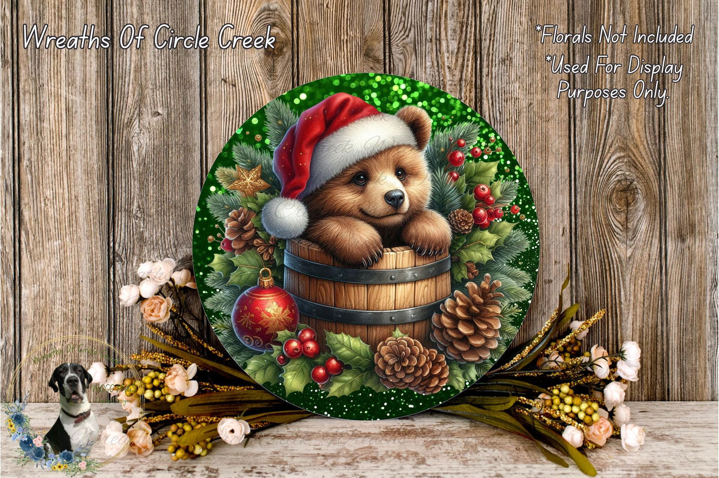 A round wreath with a bear wearing a Santa hat, surrounded by pine cones, berries, and other holiday decorations, placed on a wooden surface.