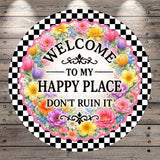 a circular sign with a floral border and a checkered border, surrounded by a wooden background. The sign displays the text "Welcome to my happy place don't ruin it" in a decorative font.