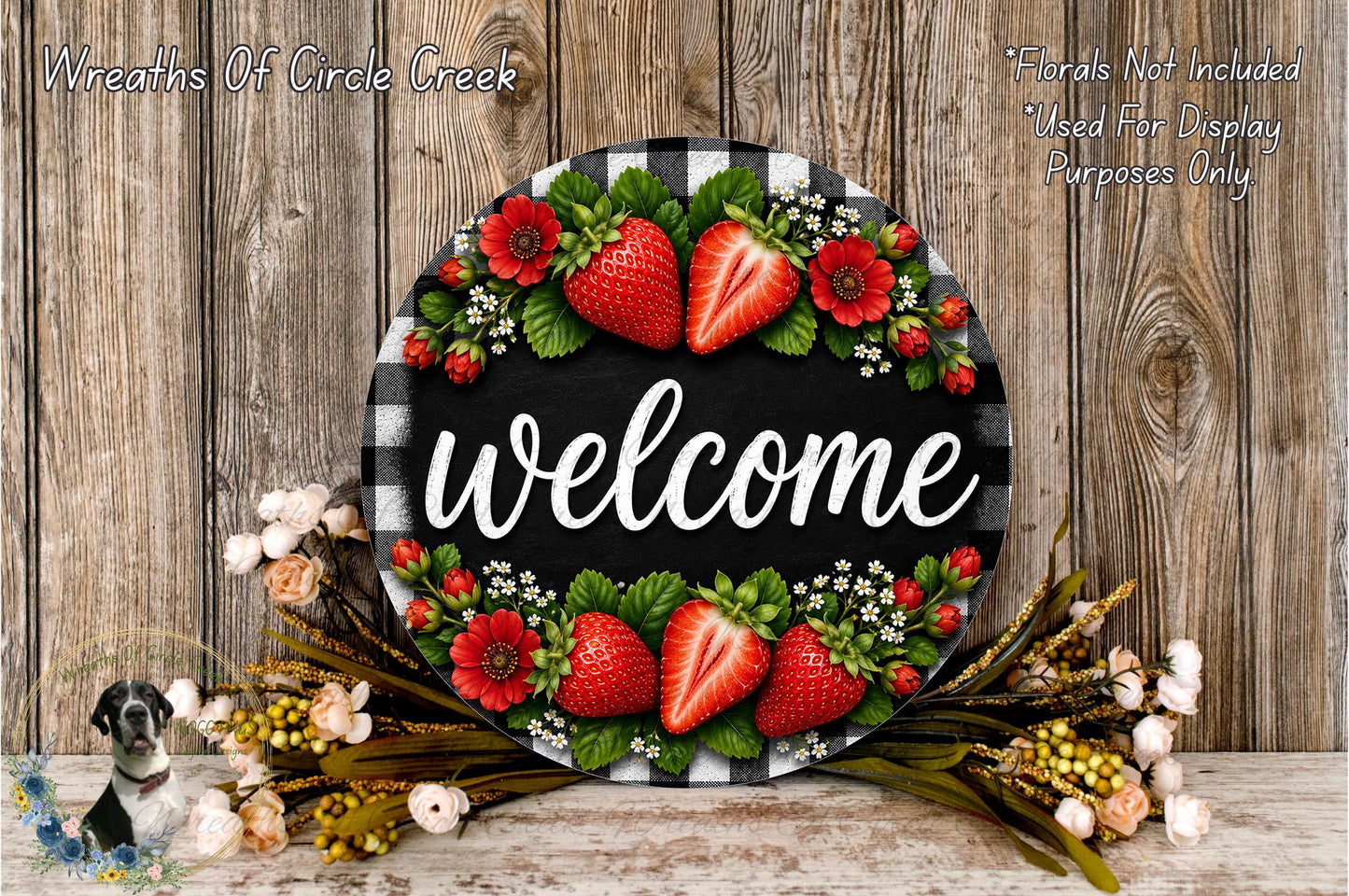 A decorative welcome sign with a black and white checkered border, surrounded by a floral arrangement of red strawberries and green leaves. The sign is placed on a wooden surface, and there is a small dog in the background.