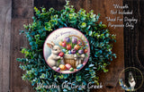 A wreath made of green foliage and featuring a white rabbit holding a basket of colorful Easter eggs and flowers.