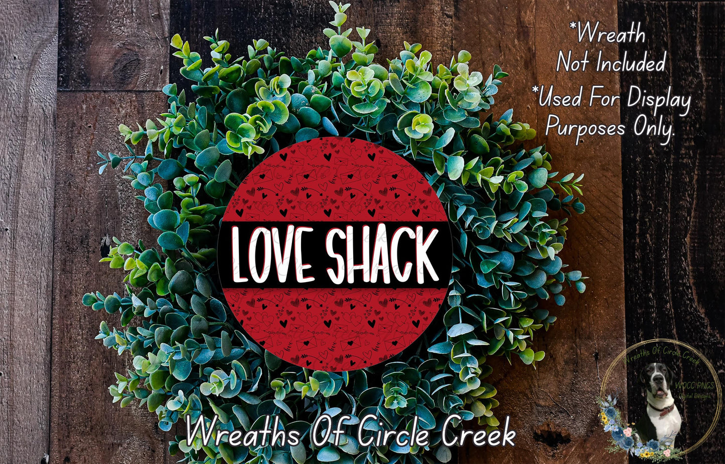 a wreath made of green leaves and a red circular logo with the text "LOVE SHACK" in the center. The wreath is displayed on a wooden surface, and there is a small dog in the bottom right corner.
