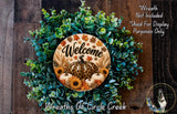 a wreath with a welcome sign, surrounded by various fall-themed elements such as pumpkins, leaves, and sunflowers. The wreath is displayed on a wooden surface, and there is a small dog in the bottom right corner of the image.