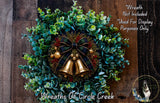 A wreath made of green leaves, red berries, and gold bells, with a plaid bow and a dog in the background.