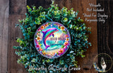 a wreath with a dolphin and the phrase "Good Vibes" on it, surrounded by a lush green plant. The wreath is displayed on a wooden surface, and there is a small dog in the bottom right corner of the image.
