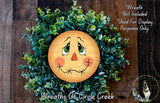 A wreath made of green plants and a yellow face with a red nose, resembling a scarecrow or scarecrow face, is displayed on a wooden surface.