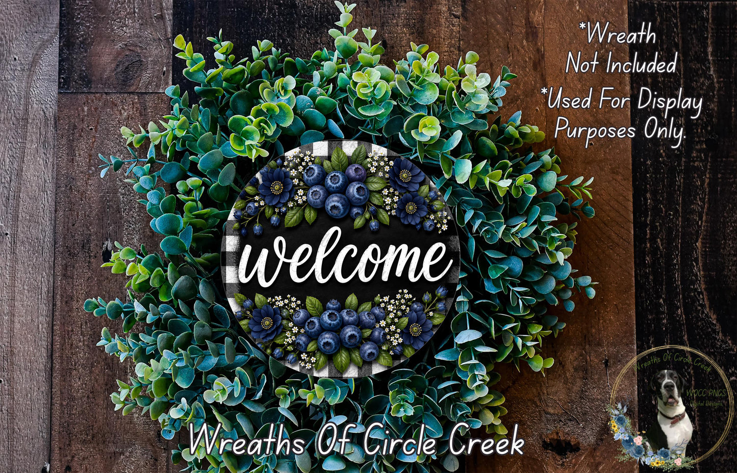 a wreath made of various green and blue foliage, with the word "Welcome" written on it. The wreath is displayed on a wooden surface, and there is a small dog in the bottom right corner of the image.