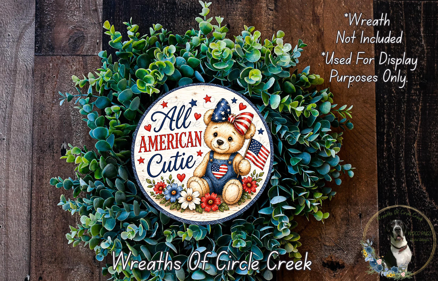 a wreath with a teddy bear wearing patriotic attire, surrounded by greenery and flowers. The wreath is displayed on a wooden surface, and there is a small dog visible in the bottom right corner of the image.