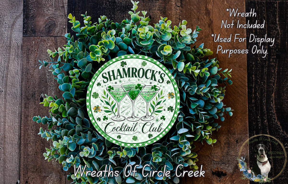 a wreath made of green leaves and flowers, with a circular emblem in the center that says " Shamrock's Cocktail Club". The wreath is displayed on a wooden surface, and there is a small dog in the bottom right corner.