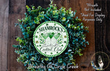 a wreath made of green leaves and flowers, with a circular emblem in the center that says " Shamrock's Cocktail Club". The wreath is displayed on a wooden surface, and there is a small dog in the bottom right corner.