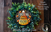 a wreath made of green foliage and candy corn, with a decorative plate in the center that says "CANDY CORN IS MY FAVORITE VEGETABLE". The wreath is displayed on a wooden surface, and there is a small dog in the bottom right corner.