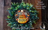 a wreath made of green foliage and candy corn, with a decorative plate in the center that says "CANDY CORN IS MY FAVORITE VEGETABLE". The wreath is displayed on a wooden surface, and there is a small dog in the bottom right corner.
