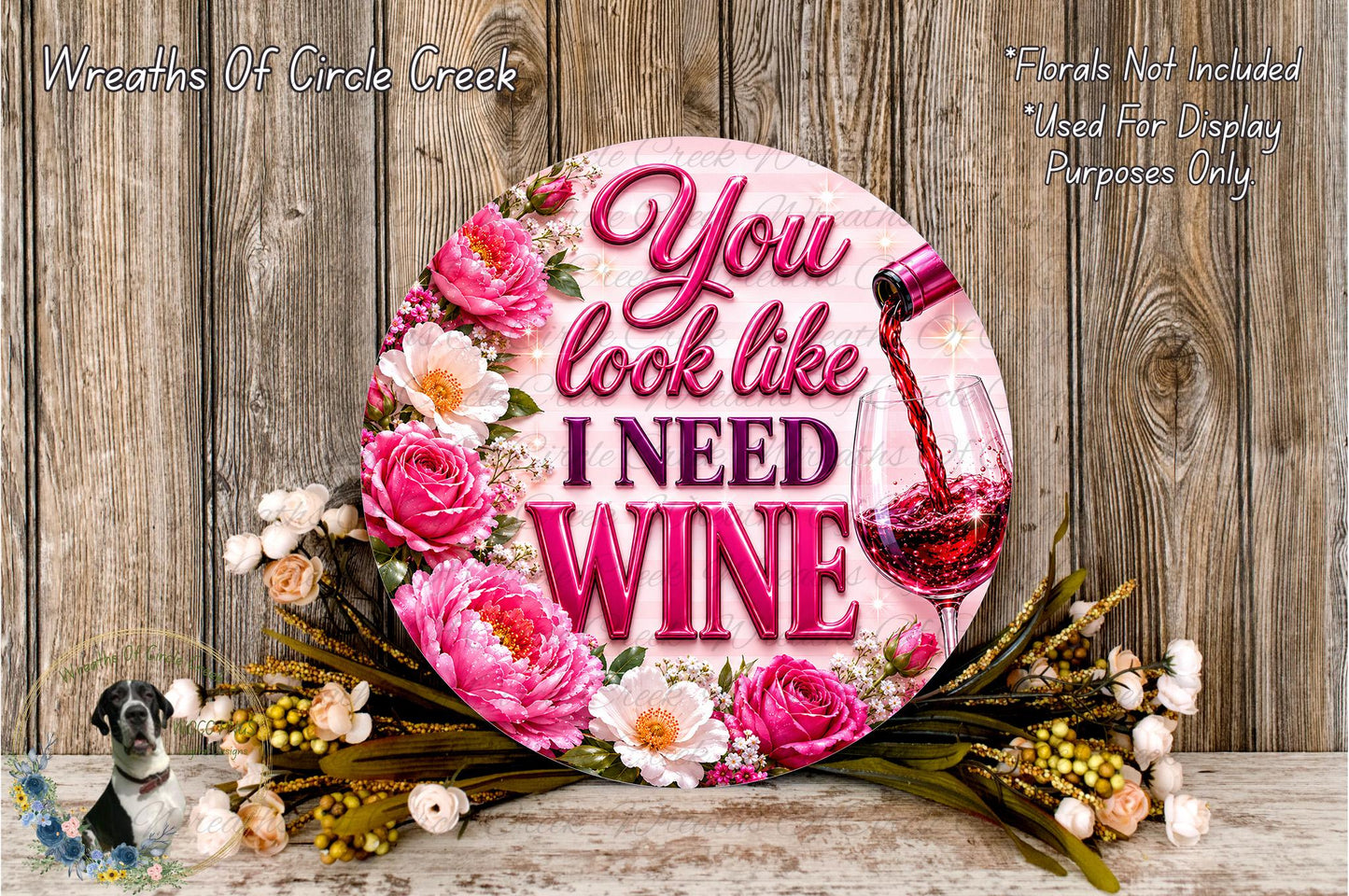 A decorative sign with the text "You look like I need wine" surrounded by pink and white flowers, with a wine glass pouring red wine. The sign is placed on a wooden surface, and there is a small bird figurine in the background.