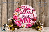 A decorative sign with the text "You look like I need wine" surrounded by pink and white flowers, with a wine glass pouring red wine. The sign is placed on a wooden surface, and there is a small bird figurine in the background.
