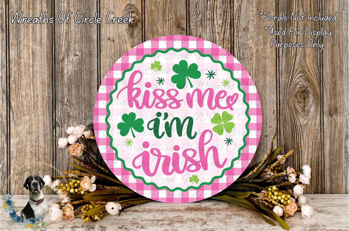 a round sign with the text "Kiss me Irish" in a pink and green checkered pattern, surrounded by a floral arrangement and a wooden background.