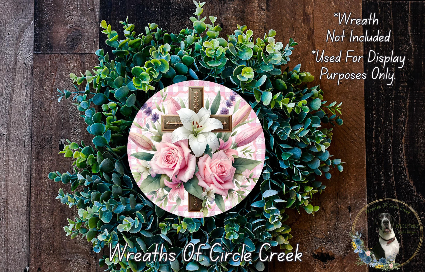 a wreath made of various green plants and pink roses, with a cross at the center. The wreath is displayed on a wooden surface, and there is a small dog in the bottom right corner of the image.