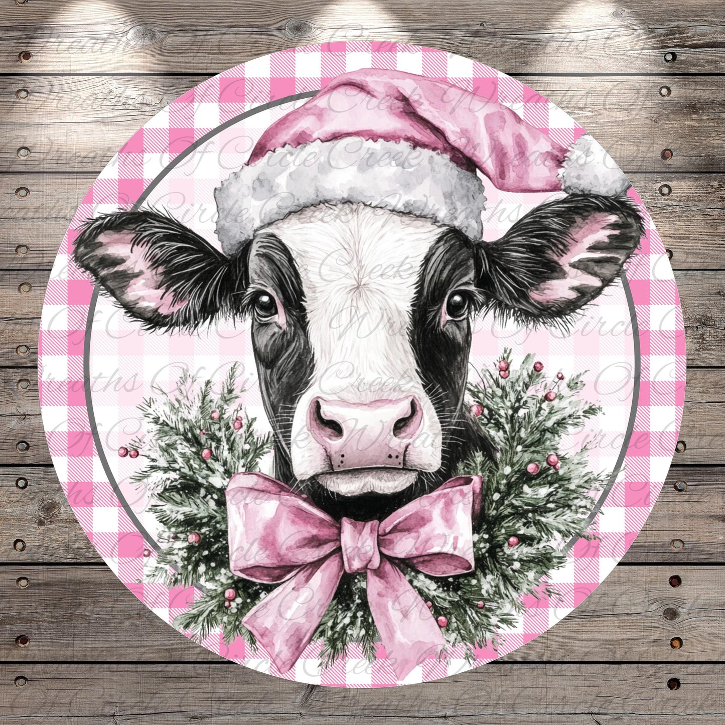 A black and white cow wearing a pink Santa hat and a pink bow tie, surrounded by a festive wreath of greenery and berries.