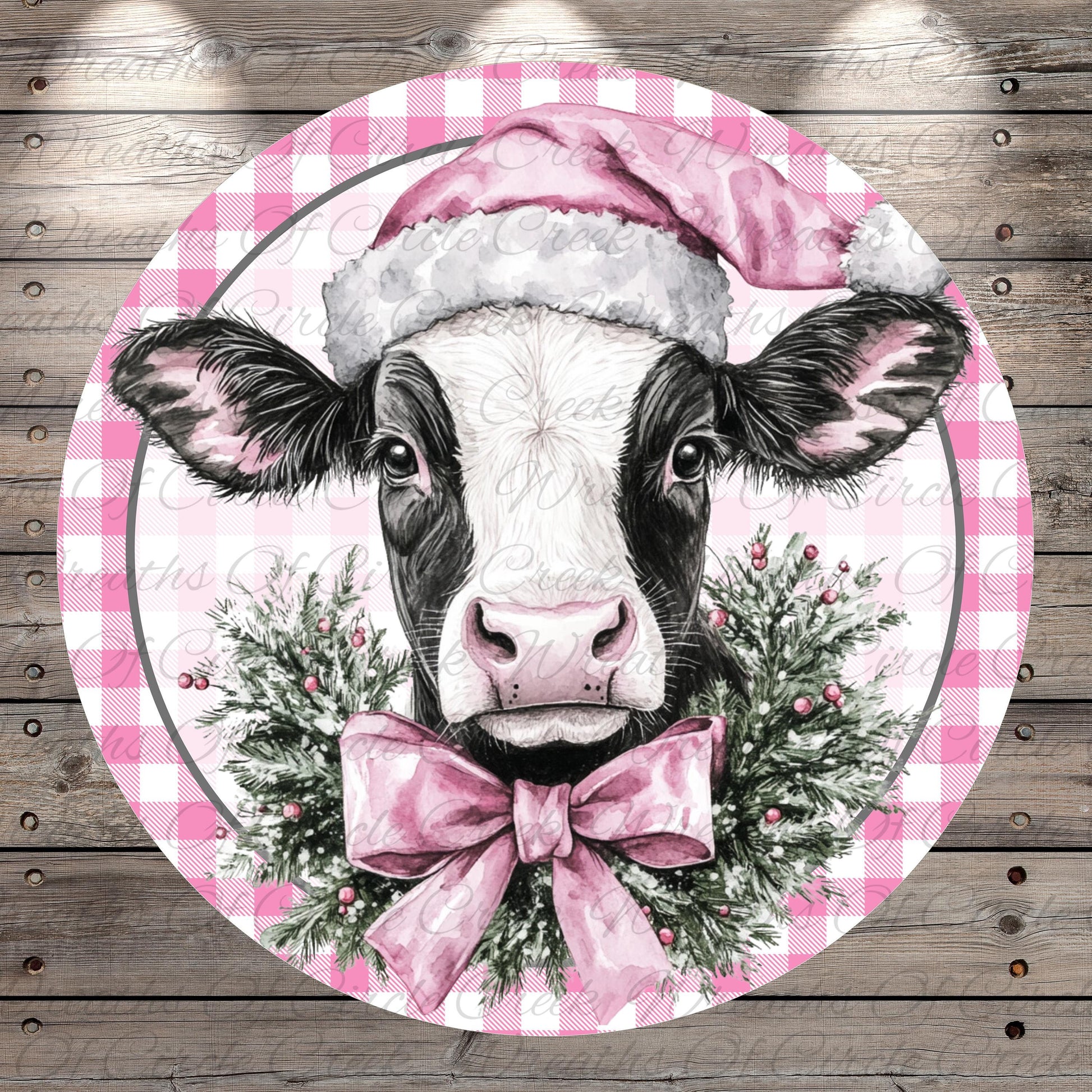 A black and white cow wearing a pink Santa hat and a pink bow tie, surrounded by a festive wreath of greenery and berries.