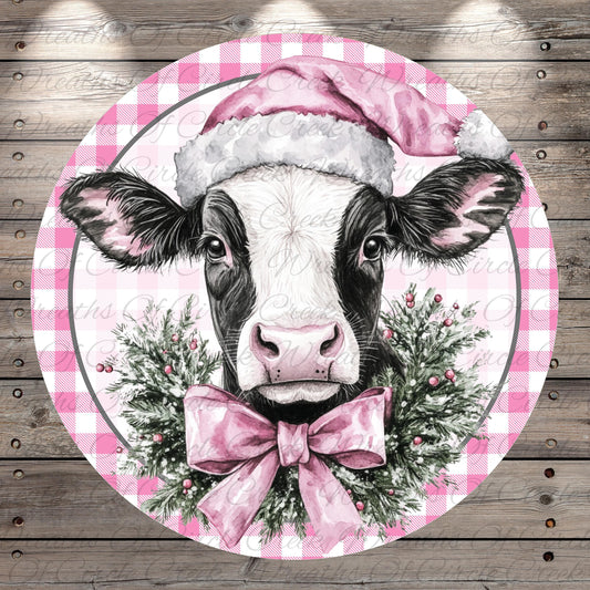 A black and white cow wearing a pink Santa hat and a pink bow tie, surrounded by a festive wreath of greenery and berries.