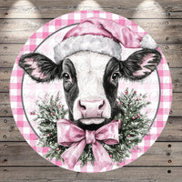 A black and white cow wearing a pink Santa hat and a pink bow tie, surrounded by a festive wreath of greenery and berries.