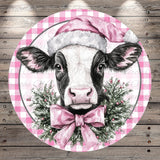 A black and white cow wearing a pink Santa hat and a pink bow tie, surrounded by a festive wreath of greenery and berries.