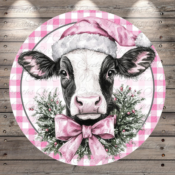 A black and white cow wearing a pink Santa hat and a pink bow tie, surrounded by a festive wreath of greenery and berries.