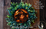 A wreath featuring a cartoon girl wearing a witch's hat and holding a pumpkin, surrounded by green foliage.