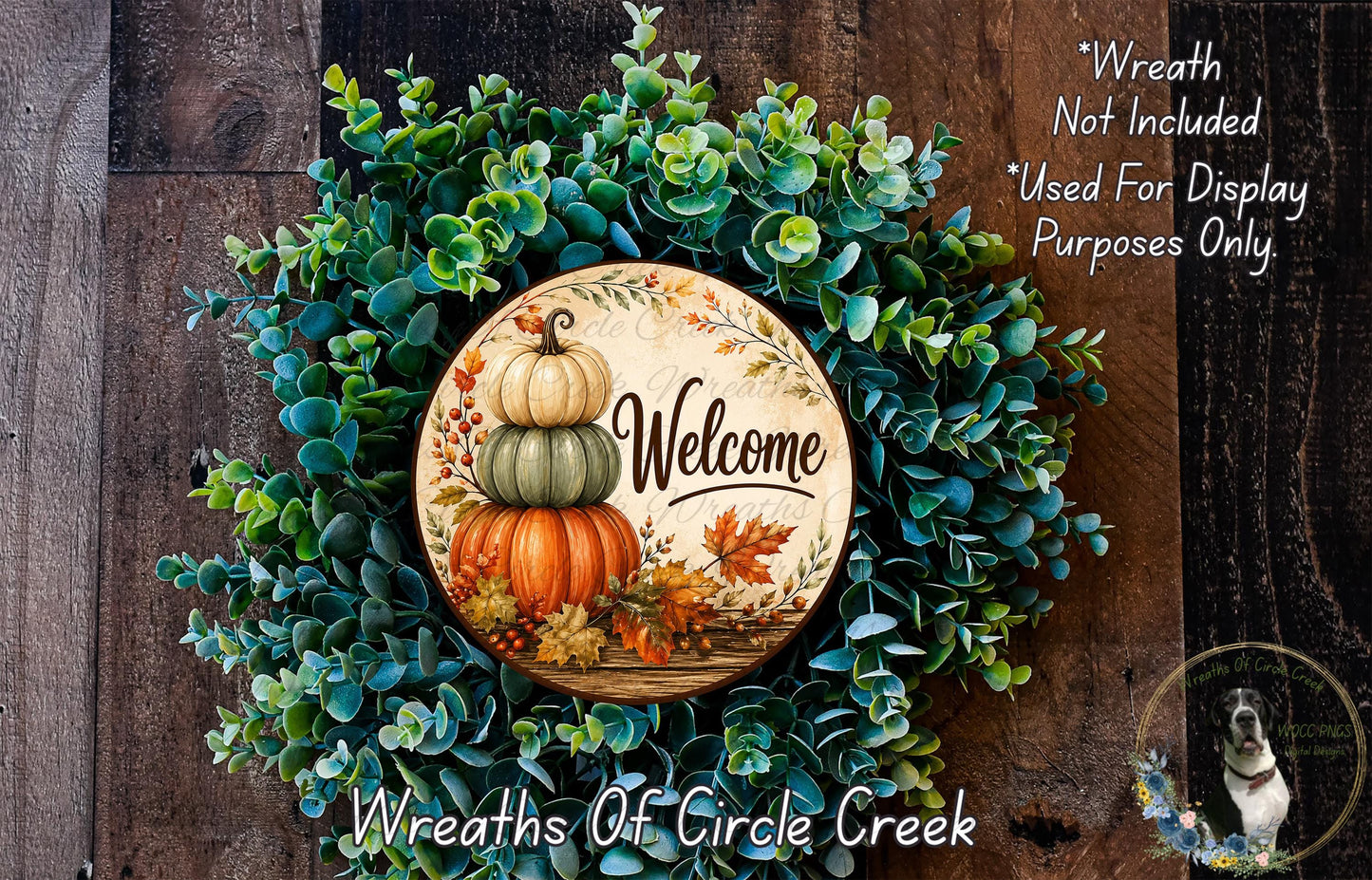 a wreath made of various green plants and a wooden sign with the word "Welcome" on it. The wreath is displayed on a wooden surface, and there is a small dog in the bottom right corner of the image.