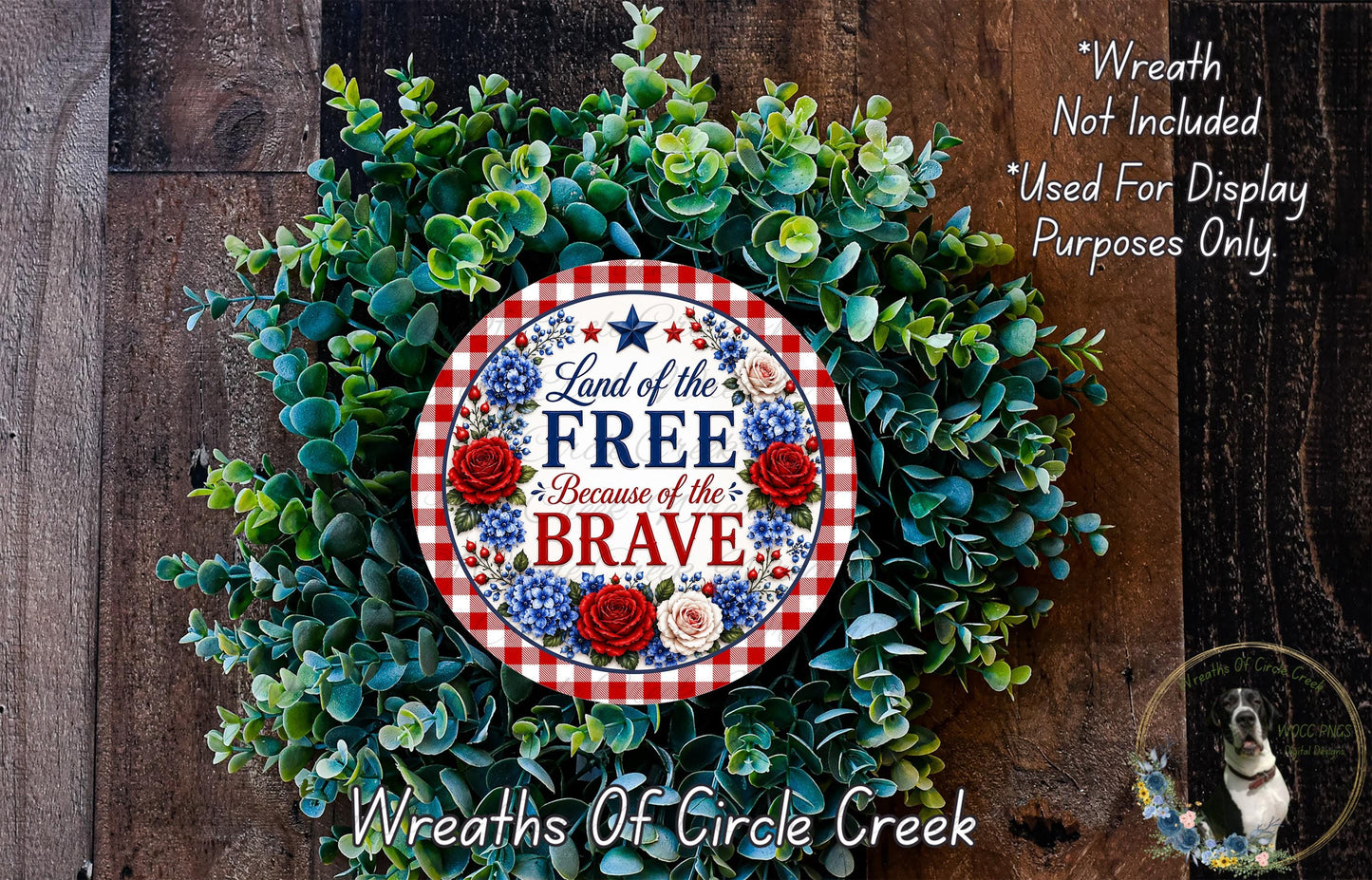 a wreath made of green leaves and flowers, with a patriotic-themed sign that reads "Land of the FREE Because of the BRAVE". The wreath is displayed on a wooden surface, and there is a small dog in the bottom right corner of the image.