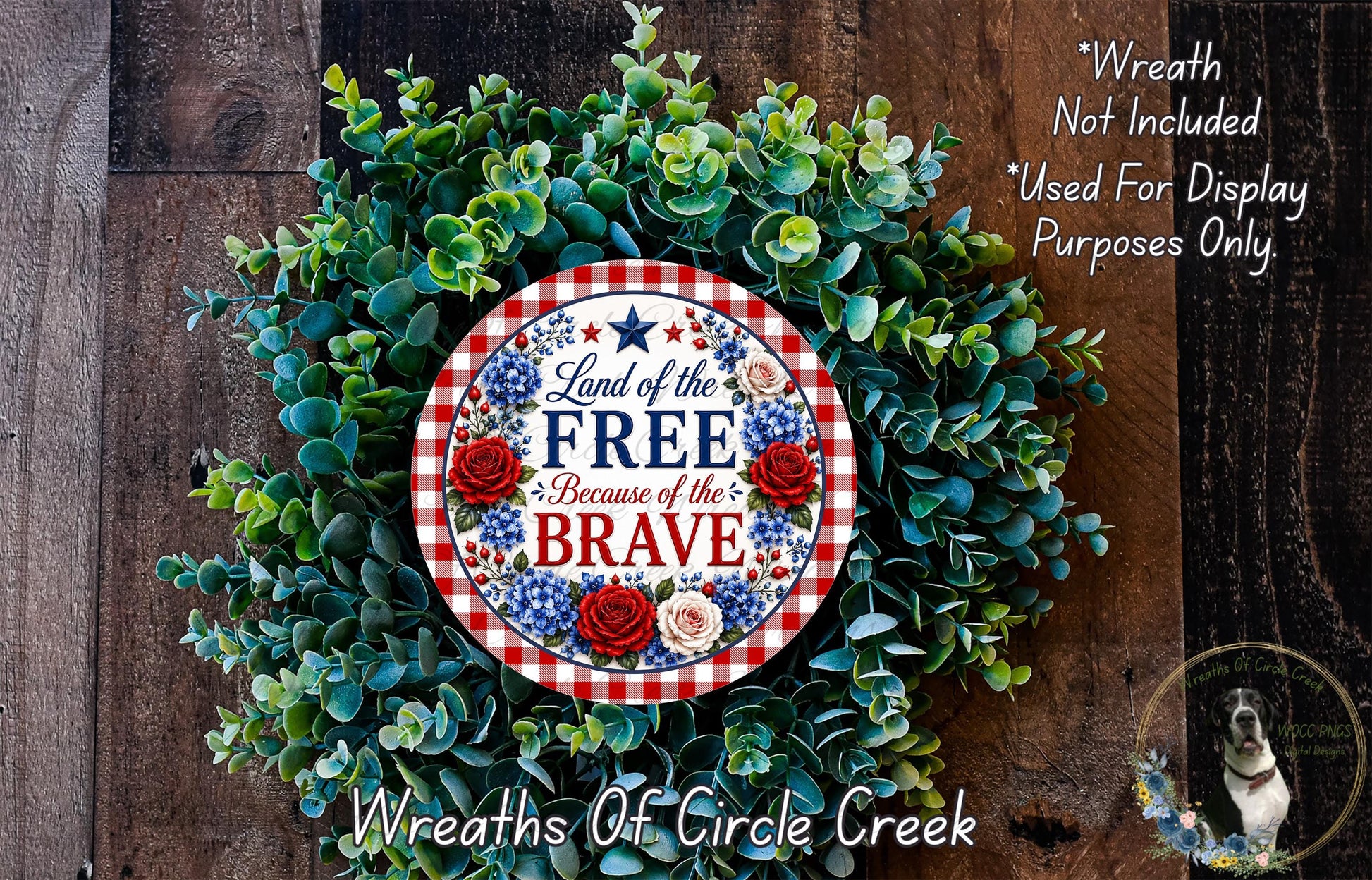 a wreath made of green leaves and flowers, with a patriotic-themed sign that reads "Land of the FREE Because of the BRAVE". The wreath is displayed on a wooden surface, and there is a small dog in the bottom right corner of the image.