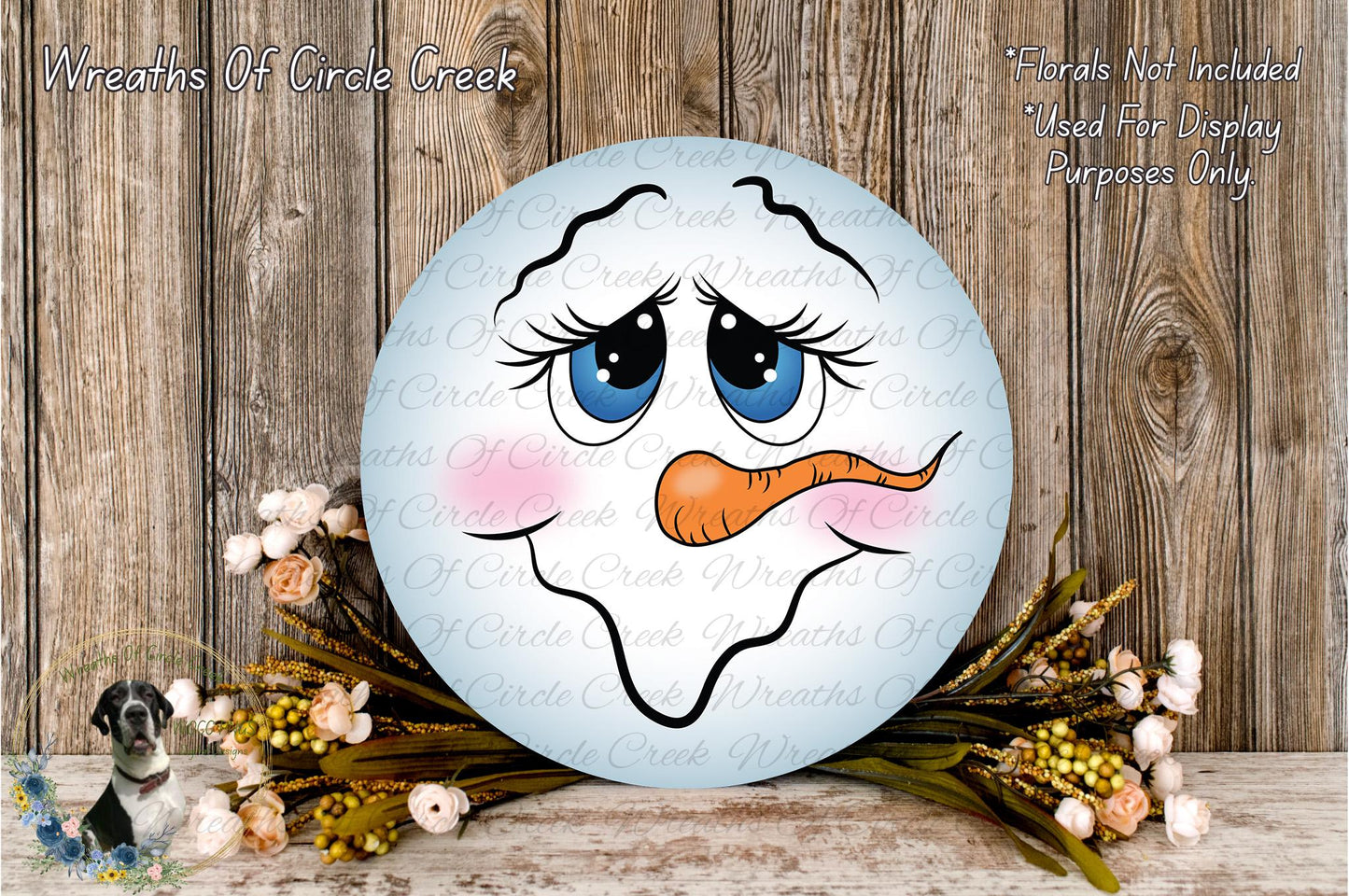 A snowman-themed wreath with a face and carrot nose, surrounded by flowers and placed on a wooden surface.